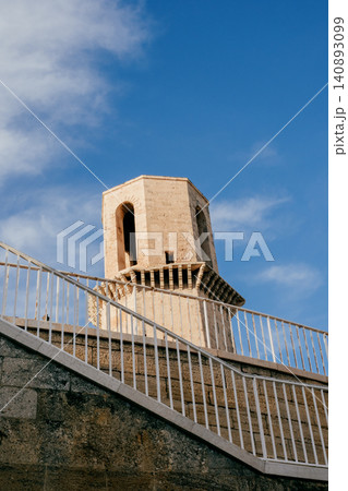 Historic stone tower against blue sky behind modern white fence architecture Historic stone tower against blue sky behind modern white fence architecture 140893099