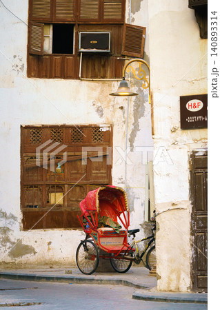 Jeddah, Saudi Arabia- Mar 08 2023, view of the wall of an Arab house with mashrabiya balconies, a red tourist bicycle stroller stands nearby, in the old city of Jeddah, Saudi Arabia 140893314