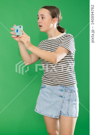 Teen elegant girl dressed in summer T-shirt and blue jeans skirt posing against green backdrop scanning barcode with smartphone or taking photo 140894762
