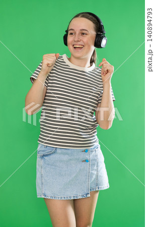 Teen elegant girl dressed in light summer T-shirt and blue jeans skirt listening to music using headphones and dancing. Female person on green background Teen elegant girl dressed in light summer T-shirt and blue jeans skirt listening to music using headphones and dancing. Female person on green background 140894793