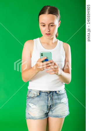 Teenage girl in denim shorts looks at smartphone screen green screen background. Chroma key compositing Teenage girl in denim shorts looks at smartphone screen green screen background. Chroma key compositing 140894905