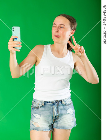 Smiling teenage girl in denim shorts takes selfie on a smartphone on green background. Chroma key compositing Smiling teenage girl in denim shorts takes selfie on a smartphone on green background. Chroma key compositing 140894936