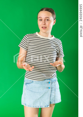 Portrait of elegant teen girl dressed in summer T-shirt and blue jeans skirt standing against green background talking with gesturing energetically and giving examples Portrait of elegant teen girl dressed in summer T-shirt and blue jeans skirt standing against green background talking with gesturing energetically and giving examples 140894995