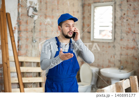 Professional worker in blue overalls speaks on a mobile phone in building under construction Professional worker in blue overalls speaks on a mobile phone in building under construction 140895177