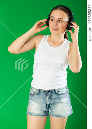 European teen girl dressed in stylish shorts listens to music using headphones and dancing. Female person on green background European teen girl dressed in stylish shorts listens to music using headphones and dancing. Female person on green background 140895220