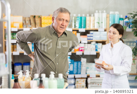Senior man visits pharmacy and suffers from backache. Girl pharmacist offer help Senior man visits pharmacy and suffers from backache. Girl pharmacist offer help 140895321