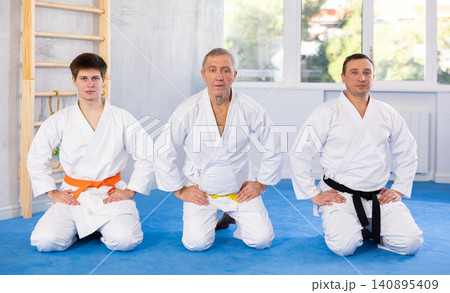 Sports team of different ages in white kimonos sit on their knees on the tatami in gym Sports team of different ages in white kimonos sit on their knees on the tatami in gym 140895409