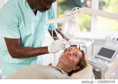 African male specialist uses ultrasonic import nozzle to care man client face skin African male specialist uses ultrasonic import nozzle to care man client face skin 140895414