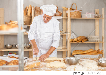 Professional elderly baker slicing dough at floured table Professional elderly baker slicing dough at floured table 140895438