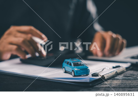 Customer signing vehicle purchase contract document for auto loan, calculating lease payment at dealership desk with blue toy car for insurance sale ownership automotive finance 140895631