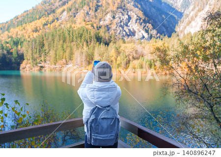 Happy woman traveler travel in jiuzhaigou national park in Sichuan, China. Tourist sightseeing and taking photo beautiful lake by mobile phone in Autumn season. Famous landmark near Chengdu, China Happy woman traveler travel in jiuzhaigou national park in Sichuan, China. Tourist sightseeing and taking photo beautiful lake by mobile phone in Autumn season. Famous landmark near Chengdu, China 140896247