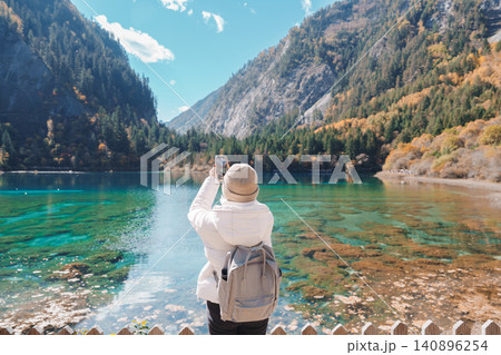 Happy woman traveler travel in jiuzhaigou national park in Sichuan, China. Tourist sightseeing and taking photo beautiful lake by mobile phone in Autumn season. Famous landmark near Chengdu, China Happy woman traveler travel in jiuzhaigou national park in Sichuan, China. Tourist sightseeing and taking photo beautiful lake by mobile phone in Autumn season. Famous landmark near Chengdu, China 140896254
