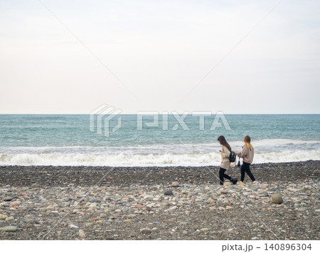 Two girls on the seashore. Looks at the waves. Walks along the coast. Off-season at the resort. Autumn in the south. Subtropics in the cold Two girls on the seashore. Looks at the waves. Walks along the coast. Off-season at the resort. Autumn in the south. Subtropics in the cold 140896304