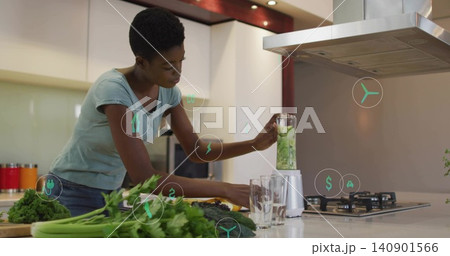 Blending woman in light blue tee preparing green drink at kitchen island, with blender and greens Blending woman in light blue tee preparing green drink at kitchen island, with blender and greens 140901566