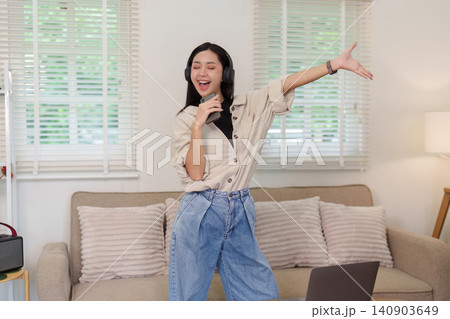 Asian woman wearing headphones singing and dancing in living room for leisure Asian woman wearing headphones singing and dancing in living room for leisure 140903649