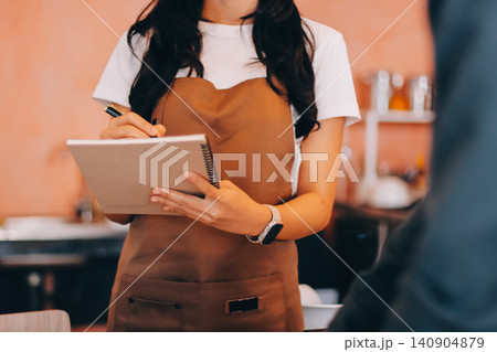 Friendly Asian female barista receives a credit card from a customer for payment. Both women are smiling in a warmly lit coffee shop with a rustic interior. Cashless payments. Small family business 140904879
