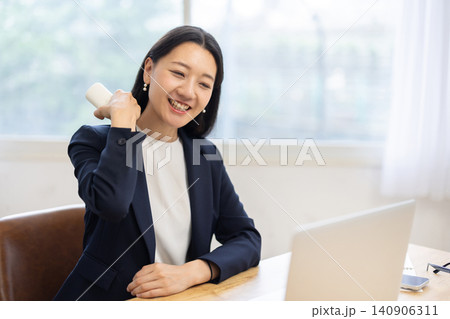 明るい窓辺のデスクでマッサージガンを使いセルフマッサージをする若い日本人ビジネスウーマン 明るい窓辺のデスクでマッサージガンを使いセルフマッサージをする若い日本人ビジネスウーマン 140906311