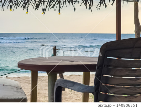 Empty table and chair at a beachfront cafe under a thatched roof with string lights and ocean view at sunset Empty table and chair at a beachfront cafe under a thatched roof with string lights and ocean view at sunset 140906551