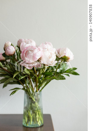 Vertical shot of lush pink peony bouquet with green leaves in ribbed glass vase on dark wooden table against neutral grey wall in natural light. 140907838