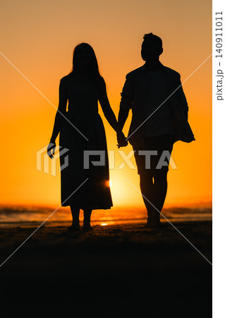 Couple walks hand in hand on the beach during sunset with orange sky and waves in the background Couple walks hand in hand on the beach during sunset with orange sky and waves in the background 140911011
