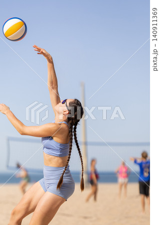 Young woman serves volleyball on sandy beach during bright day with players in background near water Young woman serves volleyball on sandy beach during bright day with players in background near water 140911369