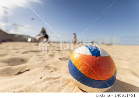 People play beach volleyball while a ball sits in the sand during a sunny day at a coastal location 140911451