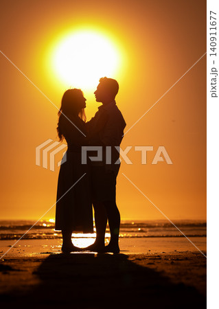 Couple stands together on the beach at sunset with the sun shining behind them 140911677
