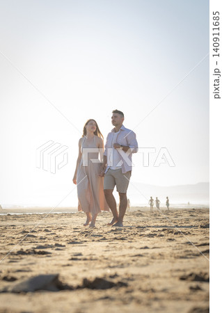 Couple walks hand in hand on a sandy beach during sunset near the ocean in a warm climate with others in the background Couple walks hand in hand on a sandy beach during sunset near the ocean in a warm climate with others in the background 140911685