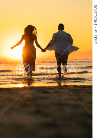 Couple runs hand in hand towards the ocean during sunset on a beach in the evening hours Couple runs hand in hand towards the ocean during sunset on a beach in the evening hours 140911738
