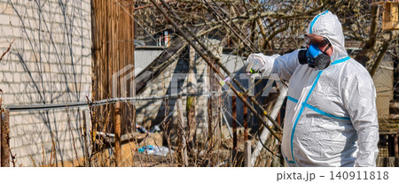 Worker in hazmat suit and full face respirator spraying fruit trees in sunny orchard, professional disinfection and pest control to protect plant health and prevent disease. Worker in hazmat suit and full face respirator spraying fruit trees in sunny orchard, professional disinfection and pest control to protect plant health and prevent disease. 140911818