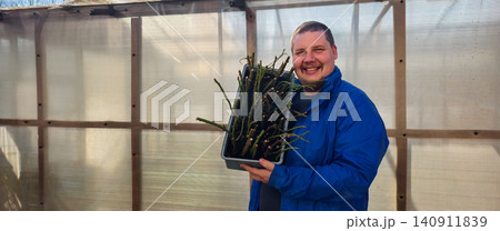 Smiling man holding a tray full of rose stem cuttings ready for propagation and planting in a greenhouse, fostering new growth and developing a horticulture hobby. Smiling man holding a tray full of rose stem cuttings ready for propagation and planting in a greenhouse, fostering new growth and developing a horticulture hobby. 140911839