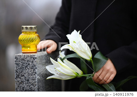 Woman holding white lilies near grey granite tombstone with candle outdoors, closeup 140911976
