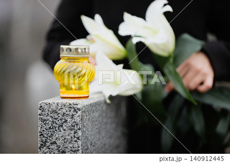 Woman holding white lilies near grey granite tombstone outdoors, focus on candle. Funeral ceremony 140912445