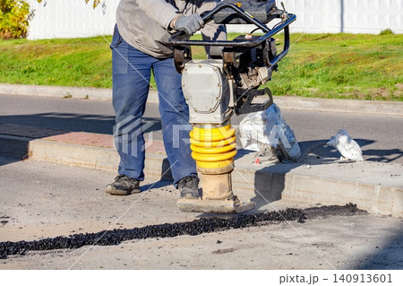 Worker operates a gas-powered tamper on a road to compact asphalt in a city street during a sunny day 140913601
