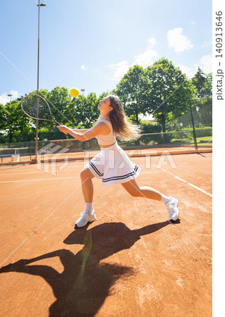 Young woman plays tennis on clay court during sunny day with clear blue sky and green trees in the background Young woman plays tennis on clay court during sunny day with clear blue sky and green trees in the background 140913646
