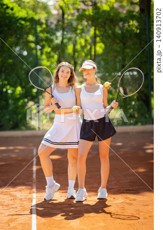 Two young women pose with tennis rackets on a sunny day at a clay court in a park Two young women pose with tennis rackets on a sunny day at a clay court in a park 140913702