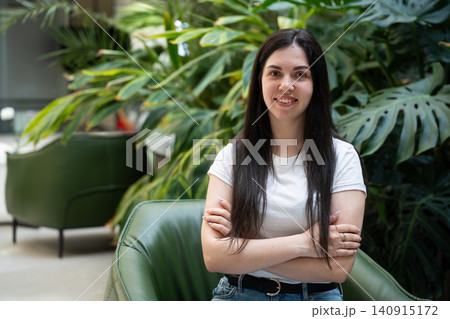 Young woman smiling with crossed arms in a modern indoor space surrounded by lush green plants. Confidence happiness lifestyle and casual portrait for personal branding 140915172
