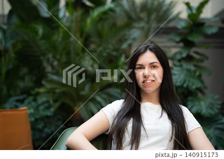 Young woman relaxing in a modern indoor space with lush green plants and smiling at the camera. Relaxation confidence lifestyle and casual portrait Young woman relaxing in a modern indoor space with lush green plants and smiling at the camera. Relaxation confidence lifestyle and casual portrait 140915173
