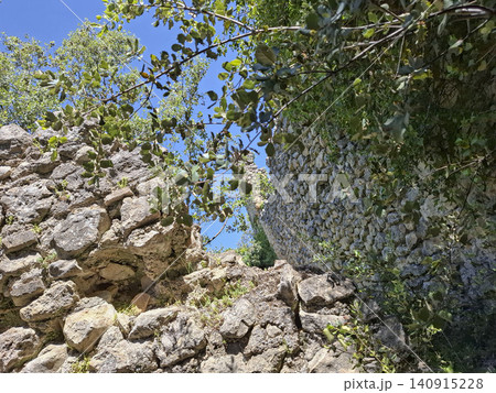 Ruins of a medieval fortress wall stand tall amid green trees in the Spanish countryside. Sunlight highlights the stone structure, revealing its ancient history. Ruins of a medieval fortress wall stand tall amid green trees in the Spanish countryside. Sunlight highlights the stone structure, revealing its ancient history. 140915228
