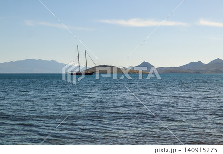 Sailing boat anchored in a tranquil bay off a distant island shore with mountains and clear blue sky Sailing boat anchored in a tranquil bay off a distant island shore with mountains and clear blue sky 140915275