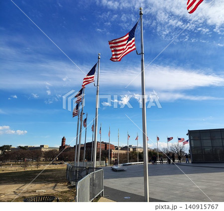 A long row of American flags flutters from tall flagpoles around the Washington Monument against a blue sky, with the occasional tourist silhouetted in the plaza A long row of American flags flutters from tall flagpoles around the Washington Monument against a blue sky, with the occasional tourist silhouetted in the plaza 140915767