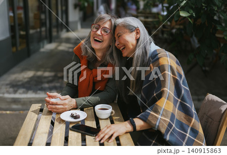 Two Diverse Mature Women Laughing Out Loud Together at an Outdoor Cafe Table with Coffee and Chocolate in an Urban Street Setting Two Diverse Mature Women Laughing Out Loud Together at an Outdoor Cafe Table with Coffee and Chocolate in an Urban Street Setting 140915863