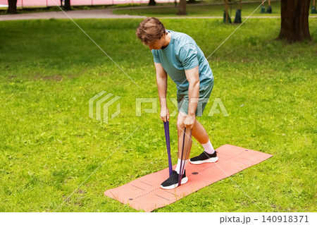 man doing an outdoor fitness workout in a park, using a resistance band on a mat for exercise and strength training. man doing an outdoor fitness workout in a park, using a resistance band on a mat for exercise and strength training. 140918371
