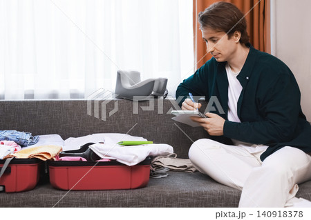 Man preparing luggage for a vacation trip, writing a packing list in a notebook. He sits on a sofa next to an open red suitcase filled with clothes. Man preparing luggage for a vacation trip, writing a packing list in a notebook. He sits on a sofa next to an open red suitcase filled with clothes. 140918378