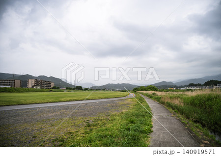 曇り空の下で緑豊かなグラウンドの横に真っ直ぐ伸びる河川敷の遊歩道と山並みの風景 曇り空の下で緑豊かなグラウンドの横に真っ直ぐ伸びる河川敷の遊歩道と山並みの風景 140919571