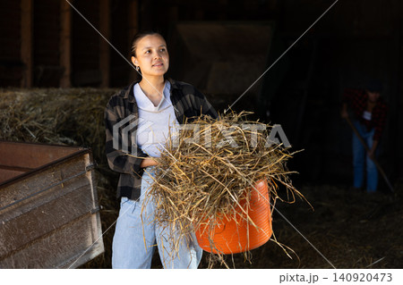 Asian young female worker walks go through corridor of stable, carrying basket with hay 140920473