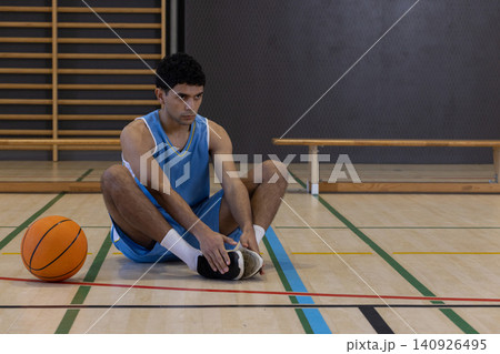 Male in blue jersey sitting on gym court reaching to hold sneaker with orange basketball nearby 140926495