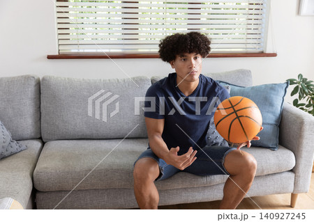 African american man sitting on sofa in living room holding basketball and gesturing African american man sitting on sofa in living room holding basketball and gesturing 140927245