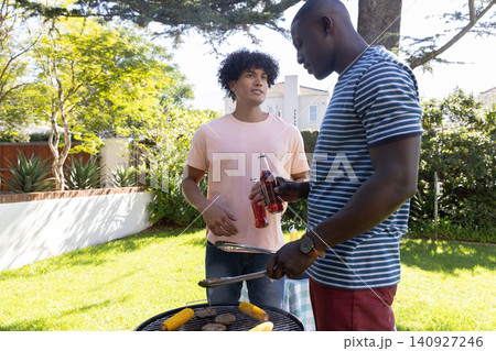 Diverse male friends grilling burgers, corn over charcoal grill holding soda bottles in backyard 140927246