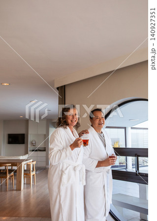 Diverse senior couple wearing bathrobes holding red drinks, gazing through round window in suite Diverse senior couple wearing bathrobes holding red drinks, gazing through round window in suite 140927351